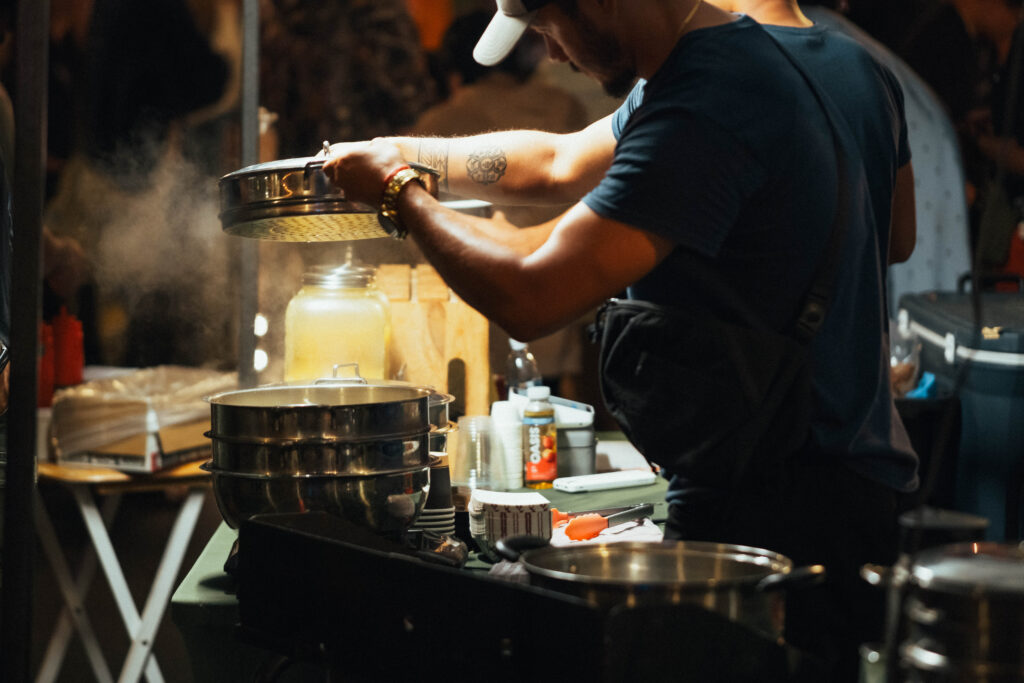 Food vendor opening a steaming pot of food in the dark of the night with twinkle lights above during the Far East Asian Night Market.