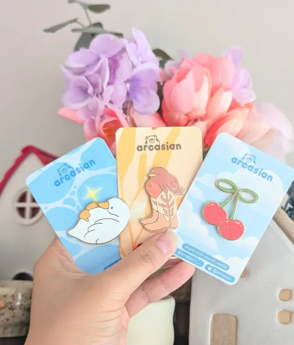 There acrylic buttons fanned out like cards. The first features a cartoon duck laying on its back, the second is a small brown bird wearing cowboy boots that are far too big and the last is two pink cherries smiling at one another. Vendor: Arcasian.