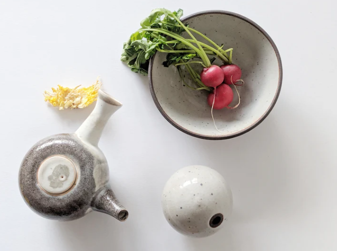 Three ceramic dishes sit upon a table. A white bowl with radishes placed inside, a white teapot and a white mug.