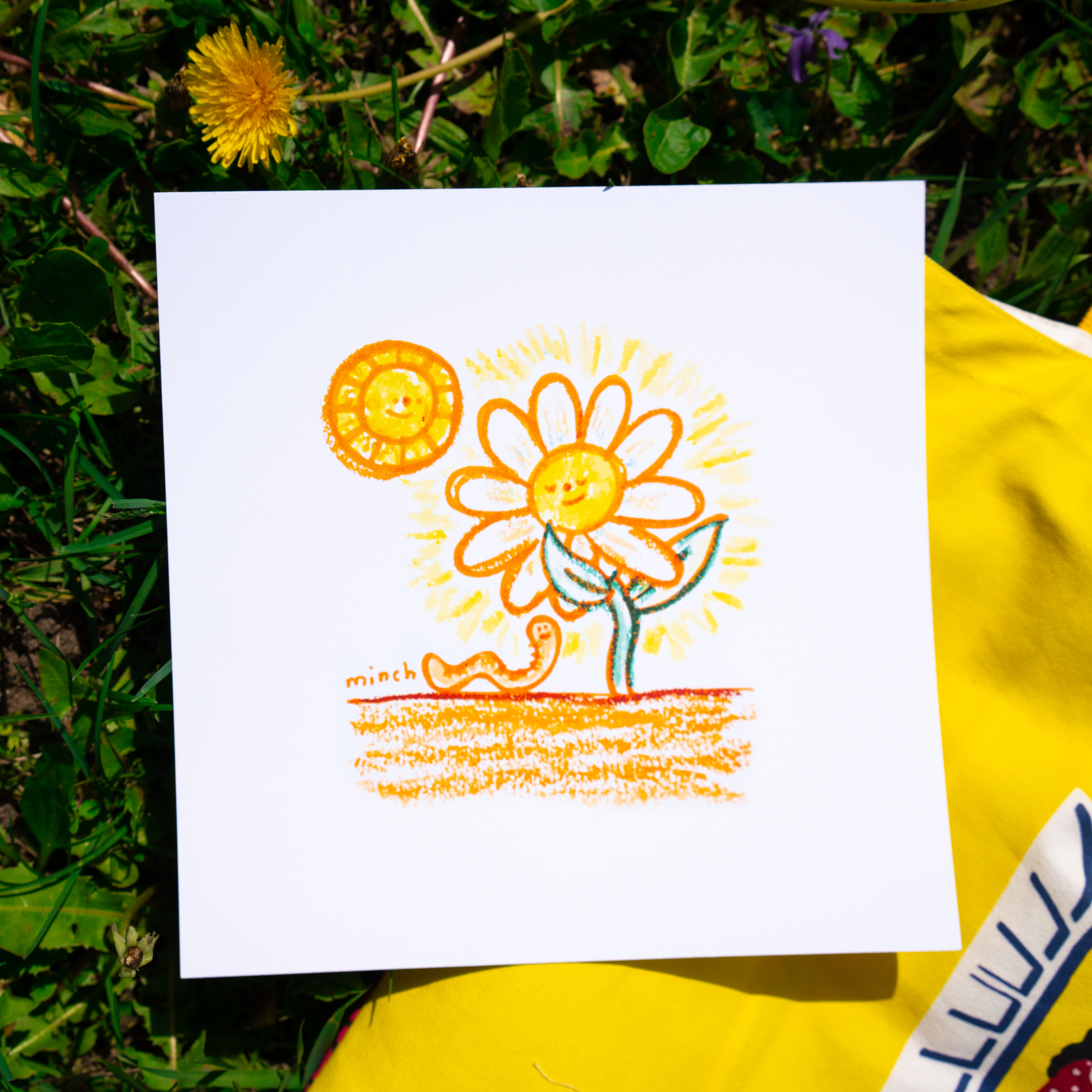 A child like drawing lays in the grass. The drawing features a sun, a flower and a worm. Vendor: Minch Creates